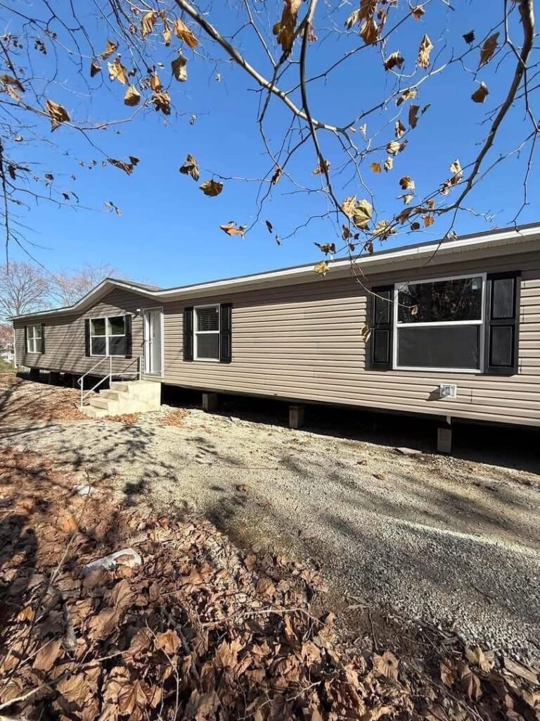 A beige mobile home with black shutters sits elevated on a gravel lot under a clear blue sky. Bare tree branches with dry leaves frame the foreground.