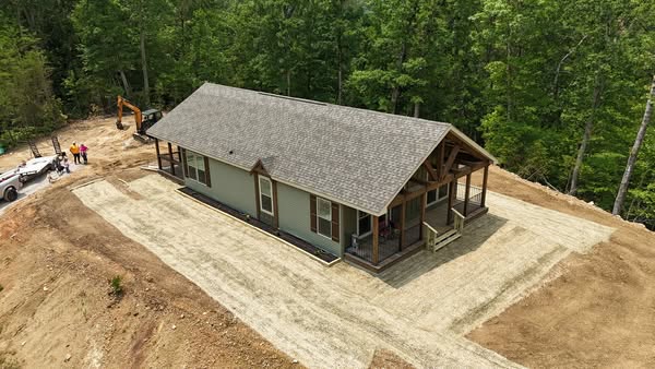 Aerial view of a single-story house with a gray roof, surrounded by dirt and trees. Construction equipment and workers are visible in the background.
