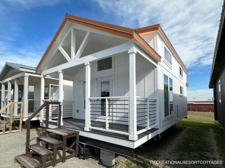 Modern tiny house with a sloped orange roof, white siding, and a front porch with railings. Clear blue sky above, conveying a calm, sunny day.