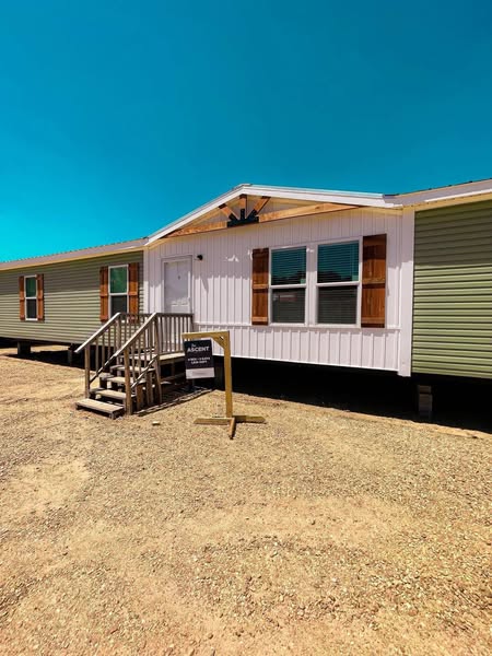 Single-story mobile home with light green and white siding, brown shutters, and a small front staircase. A sign stands nearby under a bright blue sky.