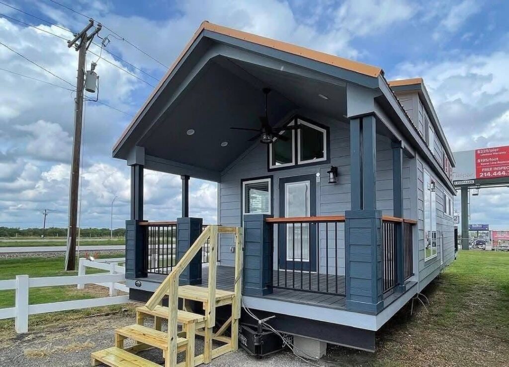 A small, modern tiny house with a gabled roof and blue-gray siding sits in a grassy area. It has a cozy porch with wooden steps, black railings, and a ceiling fan, under a partly cloudy sky.