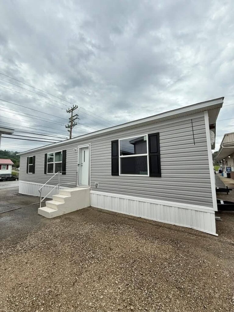 A single-story, gray mobile home with white trim and black shutters beneath an overcast sky. A small staircase leads to the front door.
