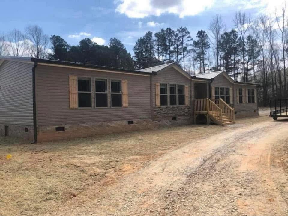A single-story, beige manufactured home with wooden shutters and a brick foundation sits on a dirt driveway, surrounded by tall trees under a partly cloudy sky.