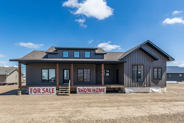 Modern single-story house with dark gray siding, large front porch, and multiple windows. Signs reading "For Sale" and "Show Home" in front, under a blue sky.
