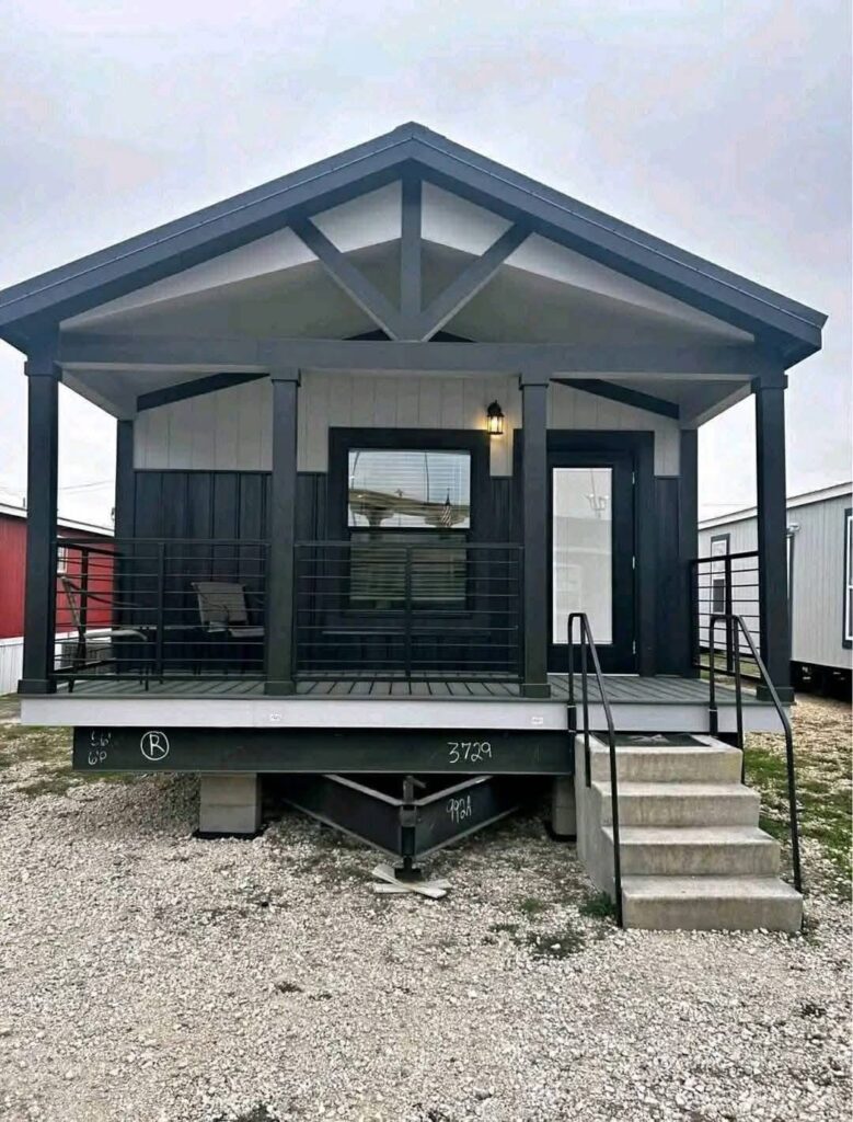 Small, modern house with gabled roof and gray siding. Features a front porch with a single chair, metal railings, and steps leading up. Overcast sky sets a calm tone.