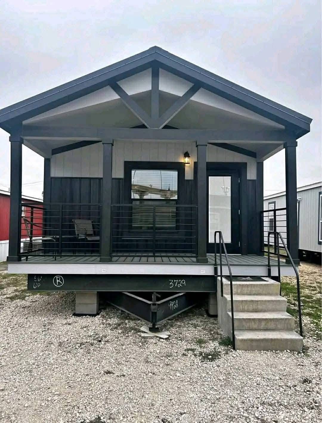 Small, modern house with gabled roof and gray siding. Features a front porch with a single chair, metal railings, and steps leading up. Overcast sky sets a calm tone.