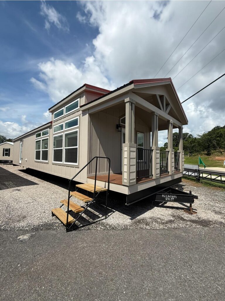 Compact mobile home with a small porch, light beige exterior, and large windows, set against a cloudy sky. The atmosphere is calm and inviting.