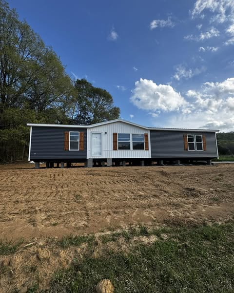 A newly built modular home with gray siding and white trim sits on a dirt foundation under a blue sky with clouds. Trees line the left side, creating a peaceful setting.