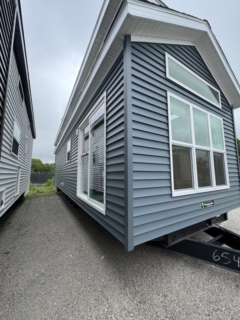 A small blue mobile home with white trim sits on gravel. Large windows on the side and front allow natural light, conveying a modern, cozy feel.