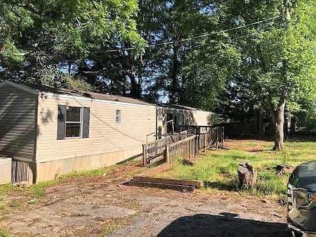 A beige mobile home with a ramp, surrounded by trees and grass, under dappled sunlight. A parked vehicle is partially visible in the foreground.