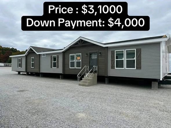A gray modular home with a gabled roof sits on a gravel lot under a cloudy sky. Overlaid text displays price details: "$3,1000" and "Down Payment: $4,000."