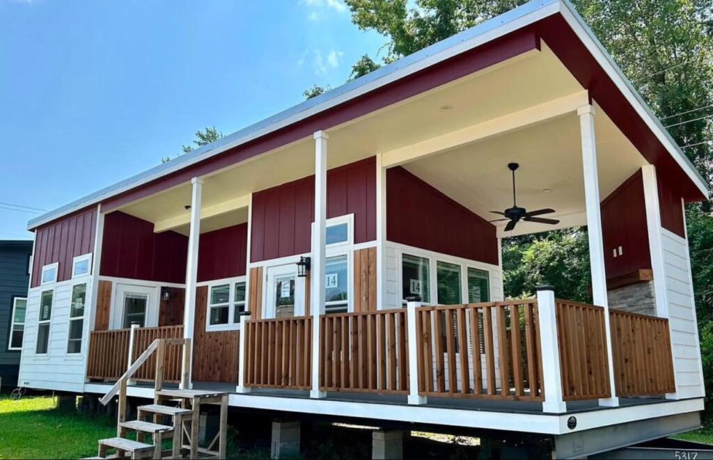 A modern tiny house with red and white exterior, large windows, and a spacious wooden porch. The setting feels bright and peaceful under a clear sky.