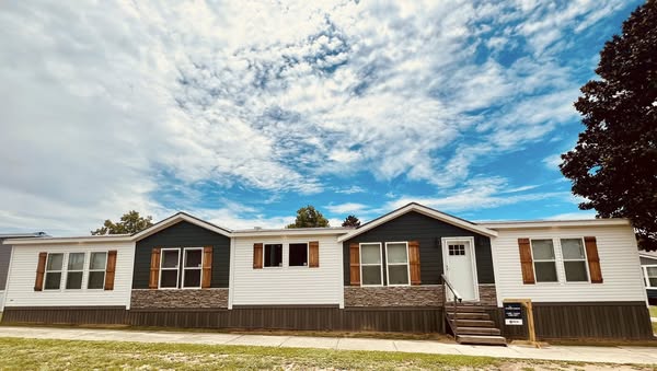 Single-story house under partly cloudy sky, featuring white siding with stone accents and brown shutters. Wooden steps lead to a white door. Calm atmosphere.