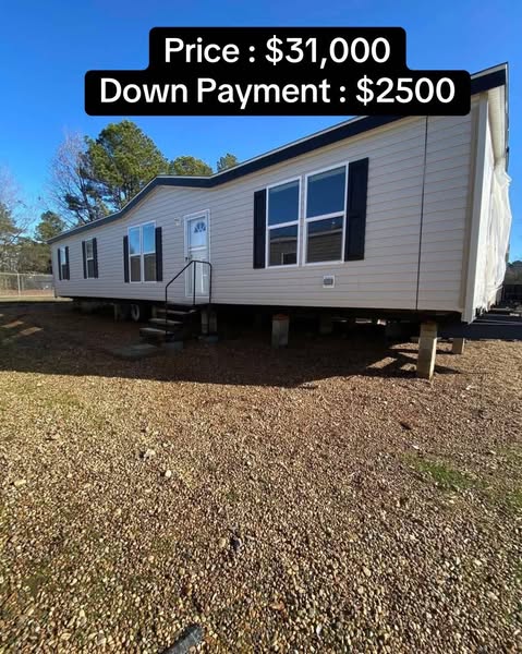A beige mobile home on a gravel lot, with large windows and a small porch. Price: $31,000, Down Payment: $2,500. Clear sky and trees in the background.