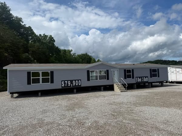 Long beige mobile home with a price tag of $79,900, featuring 5 bedrooms and 3 bathrooms. It sits on gravel with trees and cloudy skies in the background.