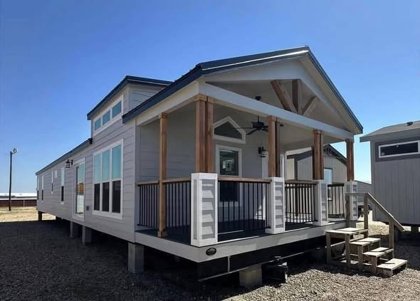 A modern tiny house on stilts with a gabled roof and front porch. Large windows line the side, and steps lead to the entrance. The scene is sunny.