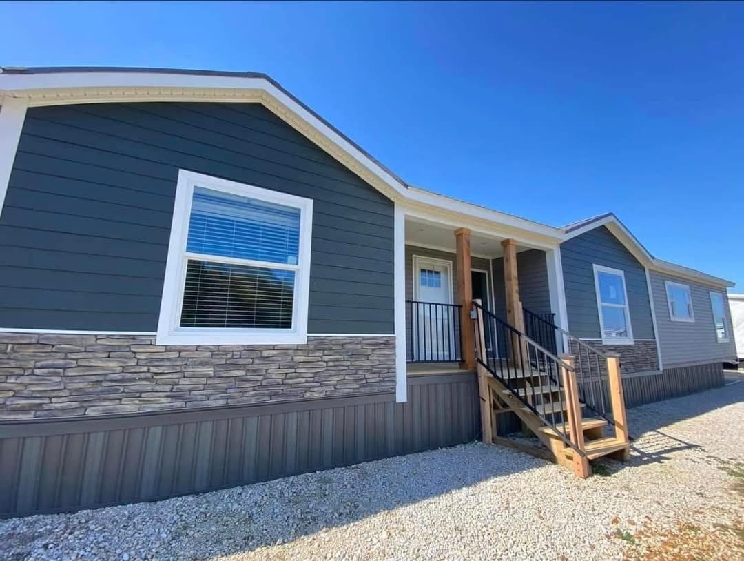 A modern mobile home with charcoal gray siding, stone accents, and white trim is shown. It has wooden steps leading to a small porch under a clear blue sky.