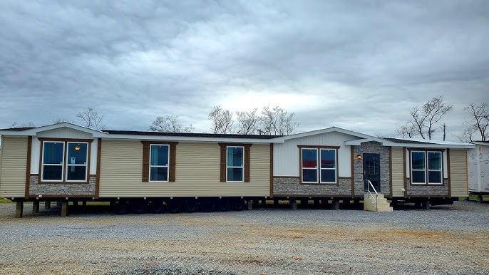 A large, modern manufactured home with a beige exterior and white trim sits on gravel under a cloudy sky. Windows feature brown shutters, enhancing its welcoming appearance.