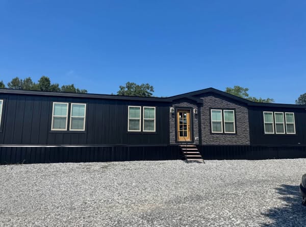 A modern, black modular home with large windows and a central glass door. The front is framed by stone accents, set against a bright blue sky.