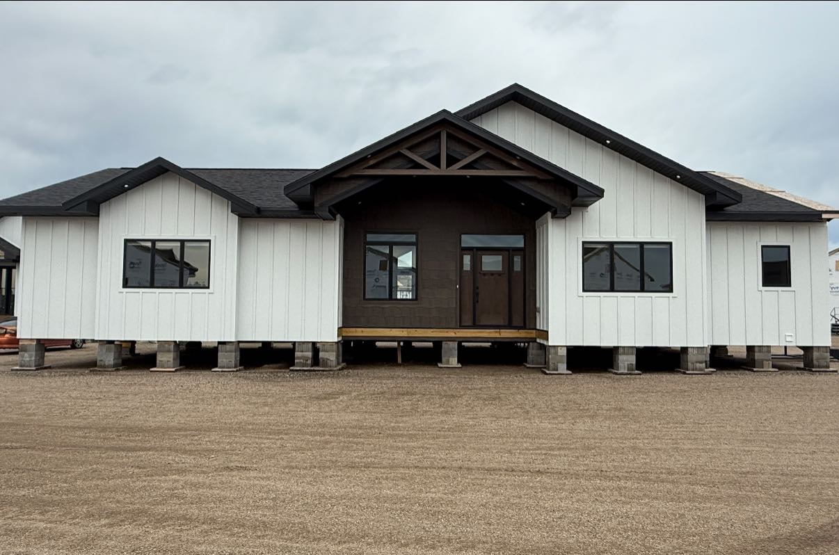 A modern white prefab house on a gravel lot under a cloudy sky. Elevated on blocks, with a dark gabled entrance and large windows.