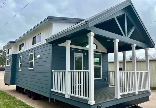 Small, modern blue tiny house with white trim and a gabled roof. It sits on a trailer, featuring a cozy front porch with railings, under an overcast sky.