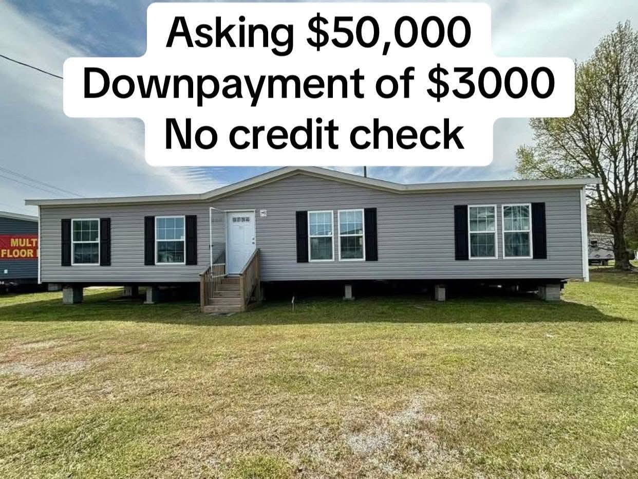 Image of a gray mobile home with white trim set against a grassy yard under a partly cloudy sky. Text above reads: "Asking $50,000. Downpayment of $3000. No credit check."