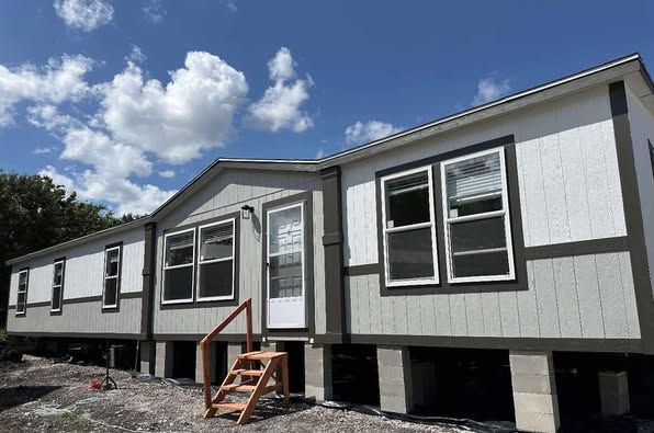 A light gray manufactured home on raised blocks under a bright blue sky with fluffy clouds. The entrance features a white door and wooden steps.