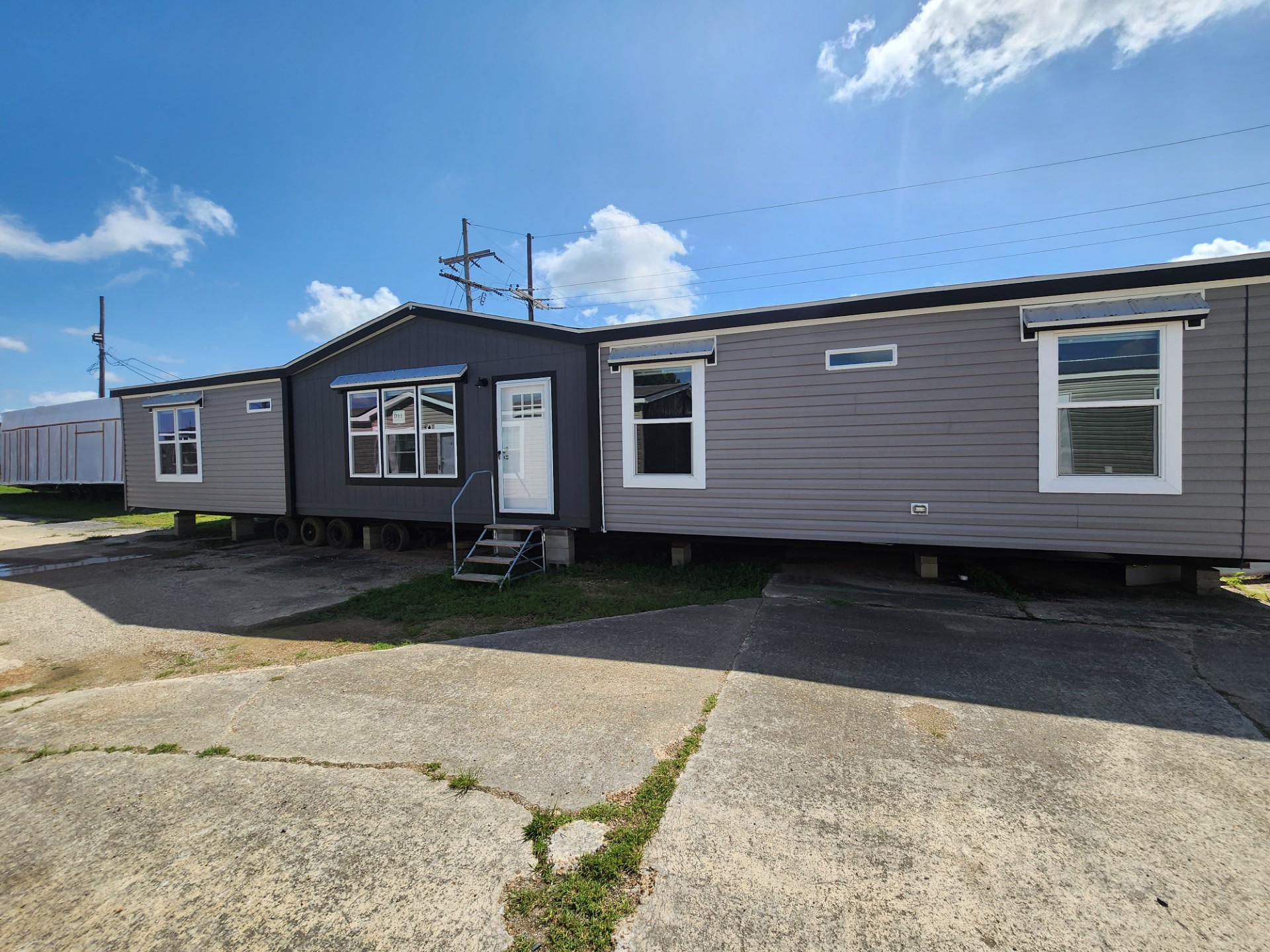 A gray modular home with a gabled roof stands under a bright blue sky with scattered clouds. Three windows and a white door are visible, set on a concrete foundation.