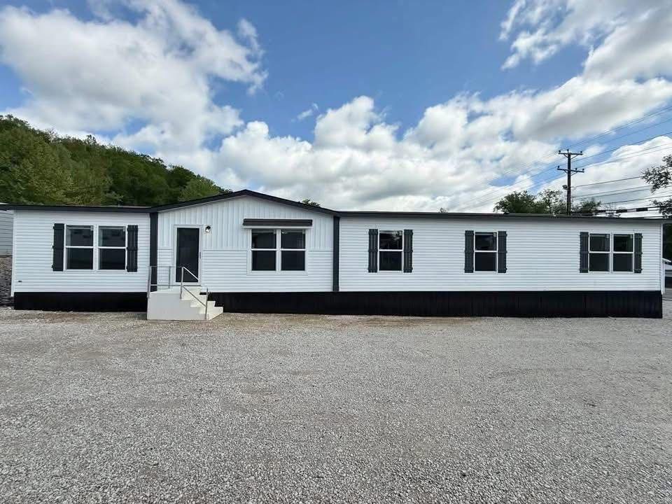A white modular home with black shutters sits on a gravel lot under a partly cloudy sky. The house has a simple porch with stairs and multiple windows.