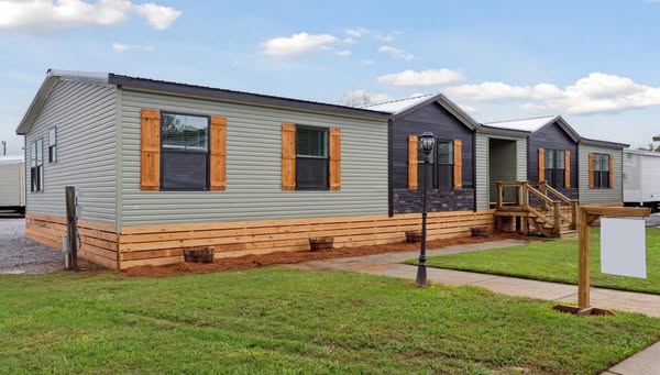 Modern single-story mobile home with light gray siding and wooden shutters. A small porch and lamp post are visible, set in a grassy yard.