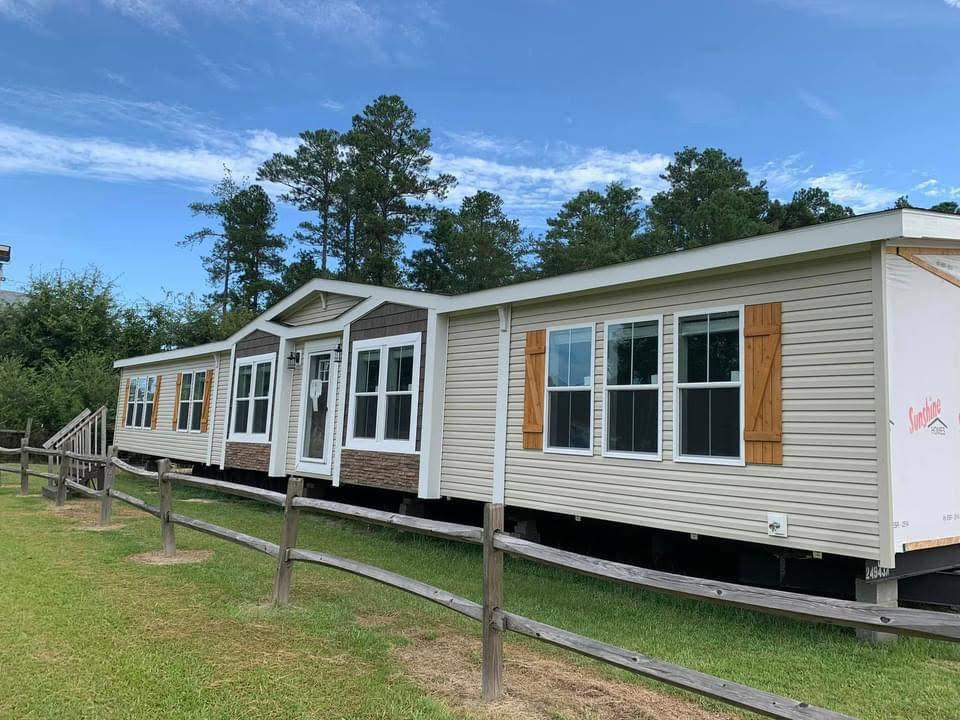 A beige modular home with large windows and brown shutters sits on a grassy lot, bordered by a wooden fence, against a backdrop of tall trees and a clear blue sky.