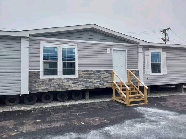 A modern, gray mobile home with white trim and stone accents, featuring a small wooden staircase leading to the front door, sits on an asphalt surface.