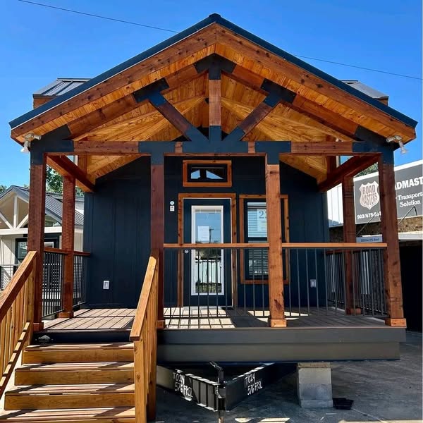Small cabin with dark walls and a wooden porch. The gabled roof features exposed beams. Stairs lead to a glass front door. Bright, clear sky.