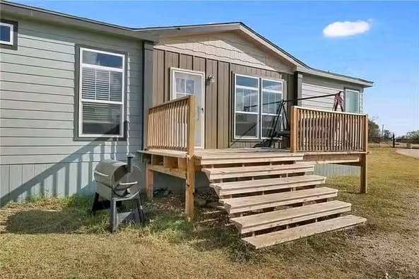 A modern mobile home with light gray siding and a small wooden porch with railings and steps. A grill is on the grassy area beside the porch.