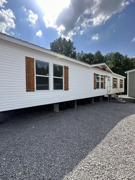 A white mobile home with wooden shutters is parked on gravel, with trees and a cloudy sky in the background. Sunlight casts shadows on the ground.