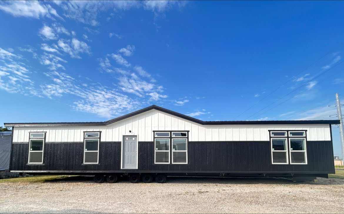 A modern mobile home with a black and white exterior under a clear blue sky. The building features a triangular roof, five large windows, and a single door.