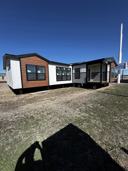 Modern modular home under clear blue sky. Structure features wooden and white siding, large windows, on a plot with dry grass, casting bold shadows.