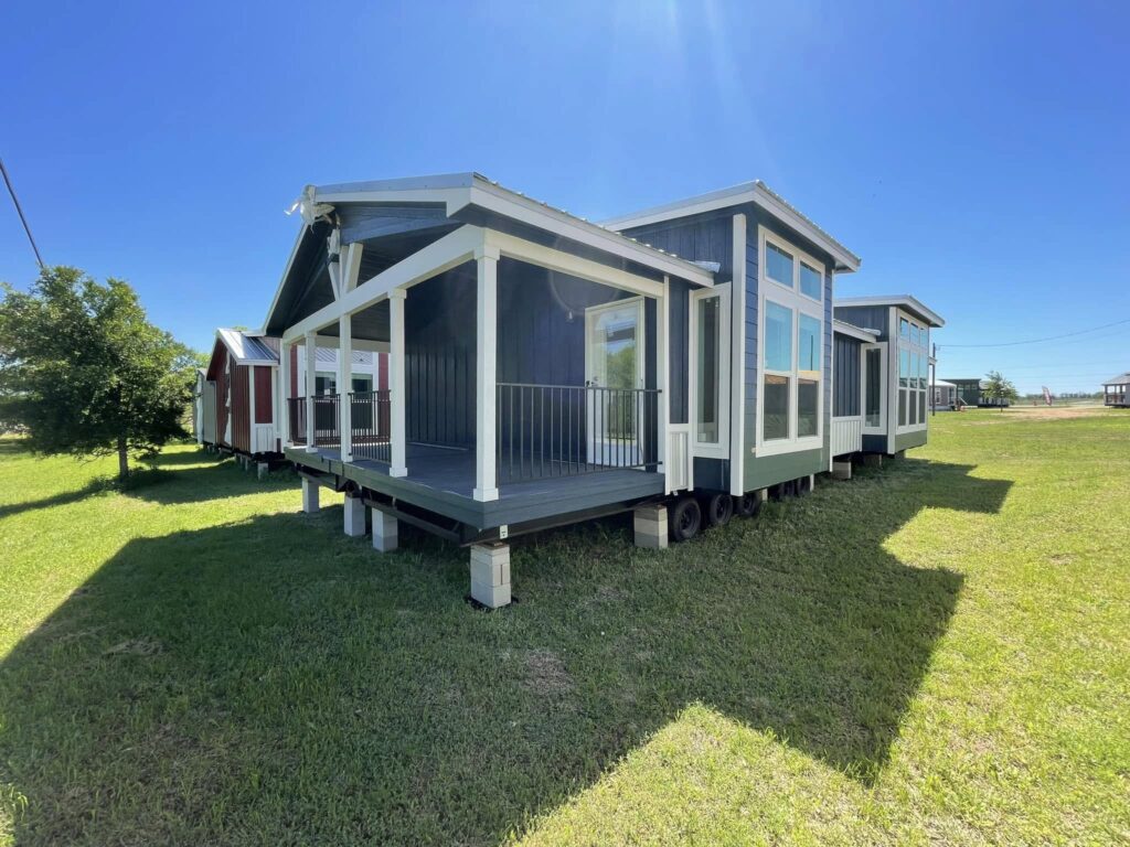Tiny house with large windows and a covered porch on a grassy field under a clear blue sky. Adjacent portable homes are visible in the background.