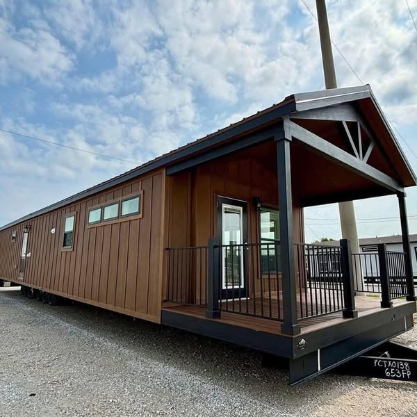 A modern, brown tiny house with a covered porch and black railing sits on gravel under a partly cloudy sky, evoking a sense of cozy simplicity.