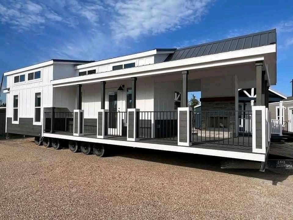 Modern white and gray tiny house on wheels with a spacious front porch and large windows. It sits on a gravel lot under a clear blue sky.