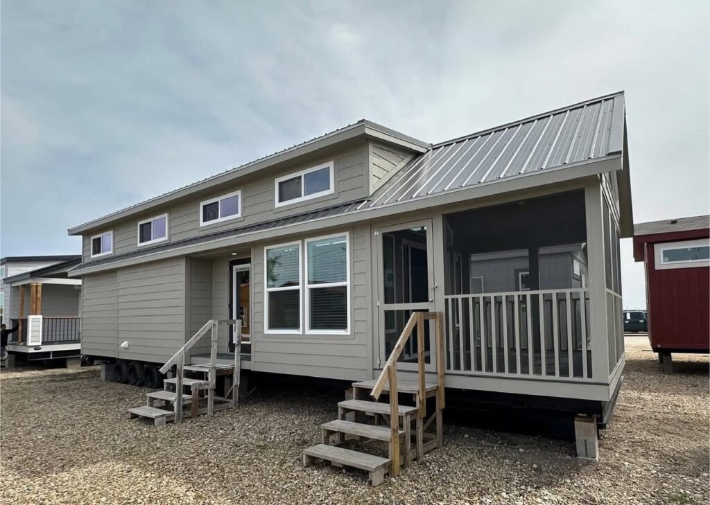 Small gray prefabricated house with a metal roof, two sets of stairs leading to entrances, and a screened porch. Overcast sky adds a muted tone.