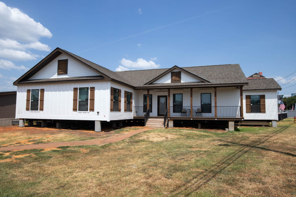 A large white house on stilts with a brown roof and wooden shutters, set on a grassy lawn under a clear blue sky with scattered clouds.