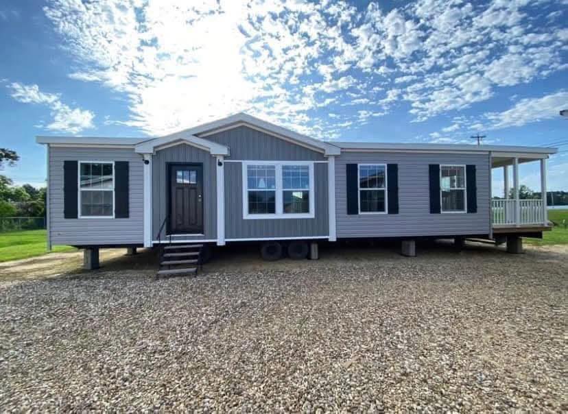A modern gray and white mobile home with black shutters sits on a gravel lot under a bright, partly cloudy sky. A small porch is visible on the right.