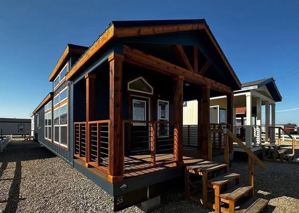 A modern tiny house with dark blue siding and wooden accents is shown. It has a front porch with rustic wood railings and steps, set against a clear blue sky.
