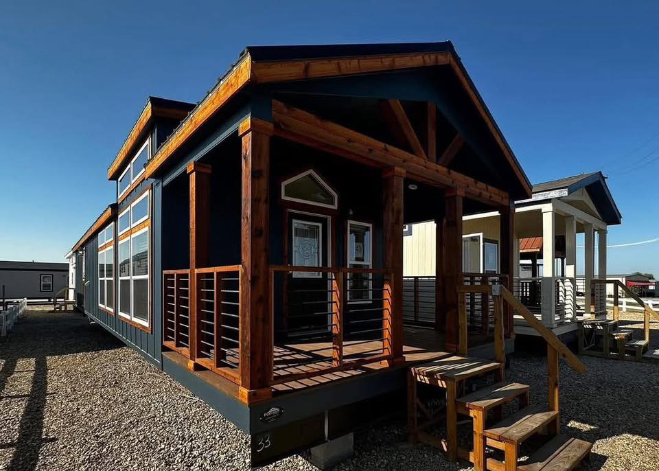 Rustic blue tiny house with wooden accents and a porch under a clear blue sky. It conveys warmth and simplicity, with a gravel pathway leading to the door.