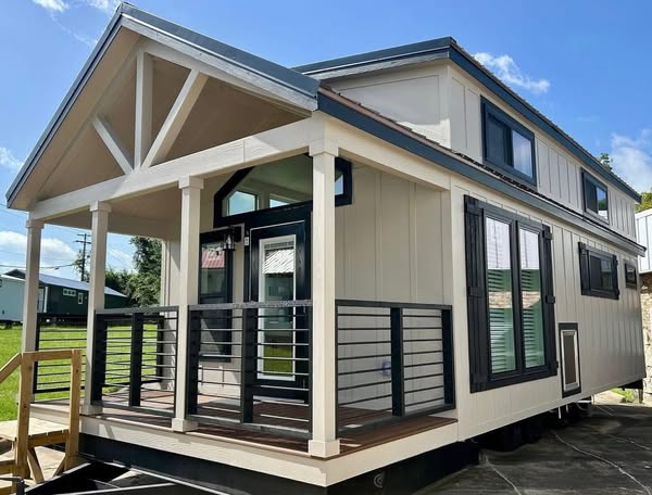 A modern tiny house with a white exterior, black trim, and large windows. The front porch features black railings and is set under a sunny blue sky.