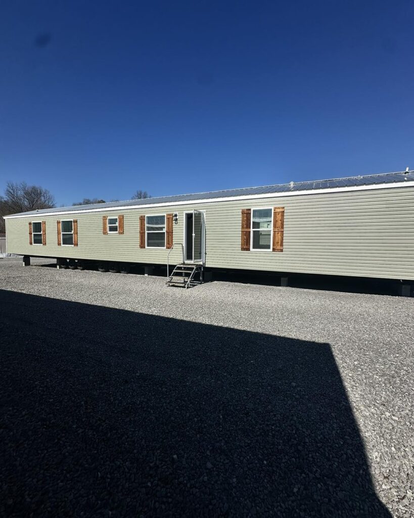 Single-story mobile home with beige siding and wooden shutters, set on gravel under a clear blue sky. A small staircase leads to the entrance.
