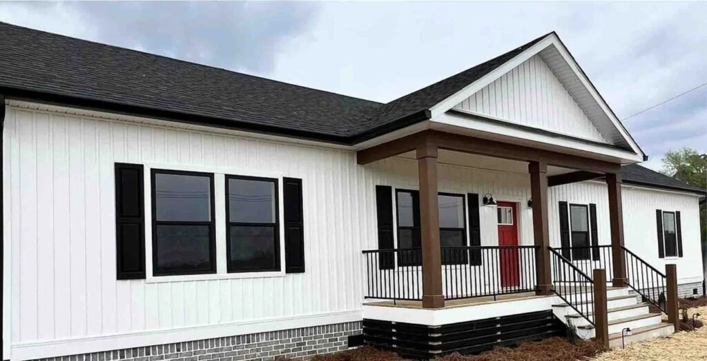 A modern white house with black shutters and a striking red door. The entrance features a wooden porch with columns, against a cloudy sky.