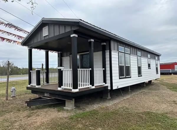 A small, modern mobile home with dark trim and a porch featuring white railing. It is set on grassy terrain under a cloudy sky, conveying a calm, rural atmosphere.