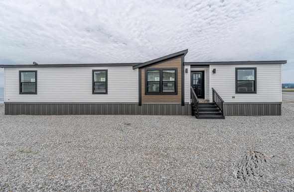 Modern manufactured home on gravel lot under cloudy sky, features white siding, a brown accent, black-framed windows, and a small front porch.